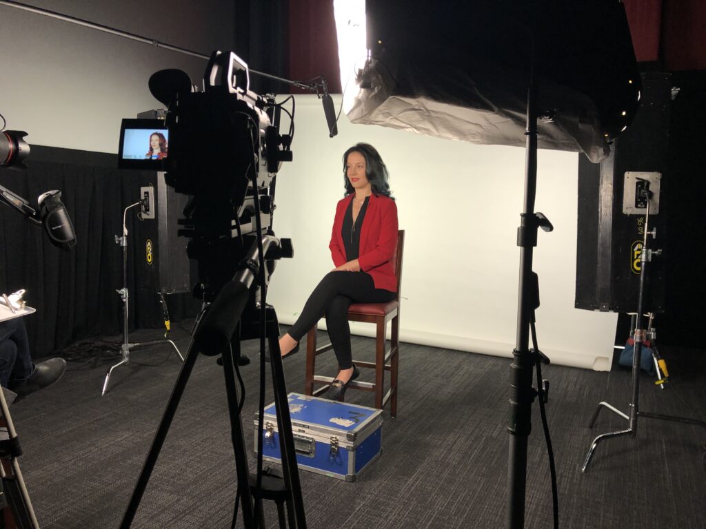 A woman with long dark hair wears a red blazer over a black shirt. Behind her is a white background. In the foreground you can see her image in the video monitor.