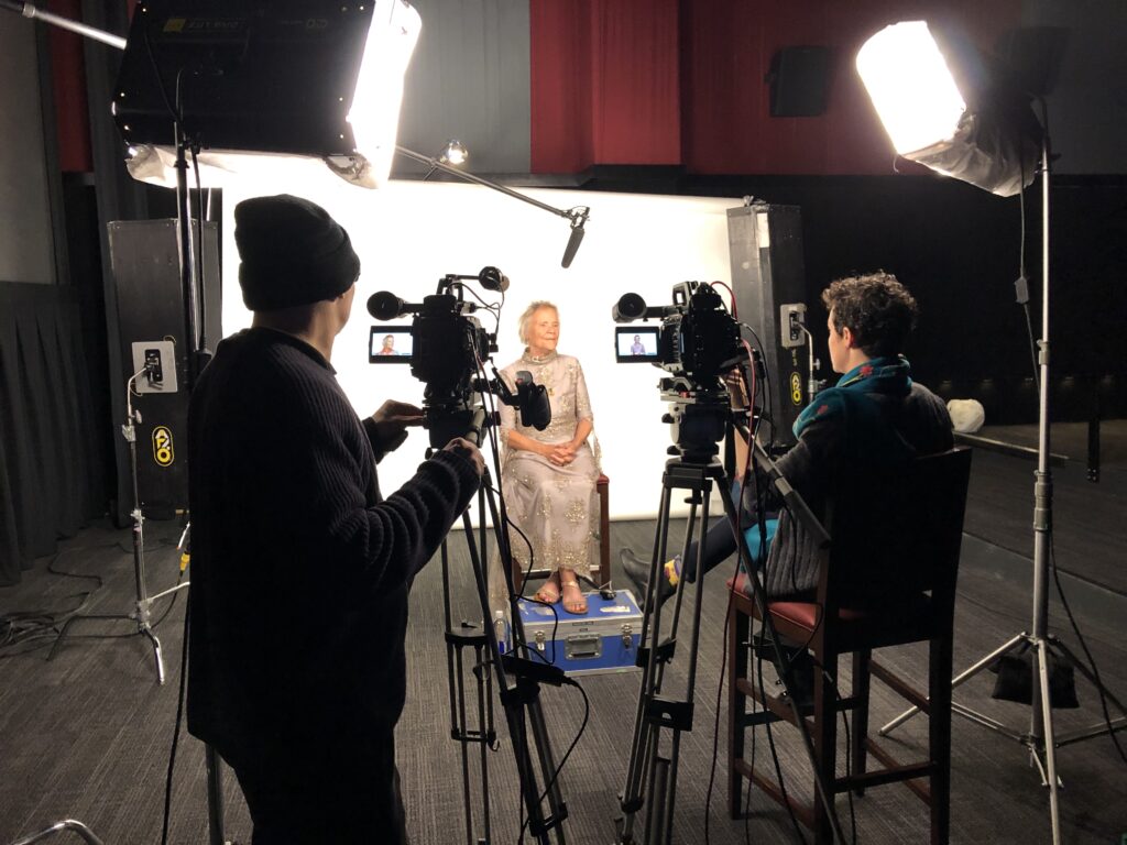 A woman in a gold dress sits in front of a white backdrop. She is surrounded by cameras and lights. In the foreground, a female director and male cinematographer prepare for the video interview.